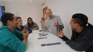 One player points towards the cards the other is holding, three other people watch, smiling. Cards are laid out on the table.